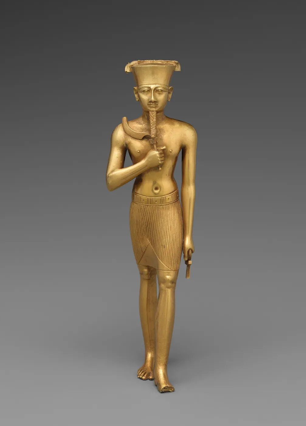 A gold statuette of Amun striding forward, wearing a flat-topped crown whose feathers are missing, holding a curved scimitar across his chest and an ankh in his lowered hand.