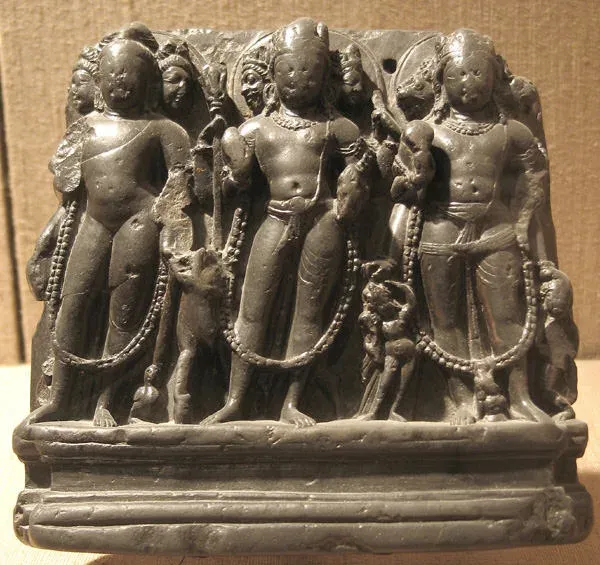 A small dark stone carving of three standing male figures side by side on a moulded base. Each wears a long garland that hangs to the knees, each has extra faces beside the central one, and small attendant figures and animals stand between them.