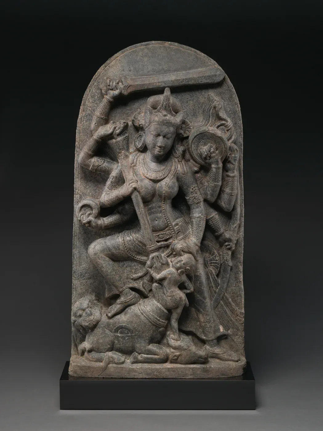 A grey stone slab with a rounded top carved in high relief. A goddess with eight arms stands with one foot on a fallen buffalo, a sword raised over her head and a shield at her side, driving a trident into a small human figure who rises from the animal's neck.
