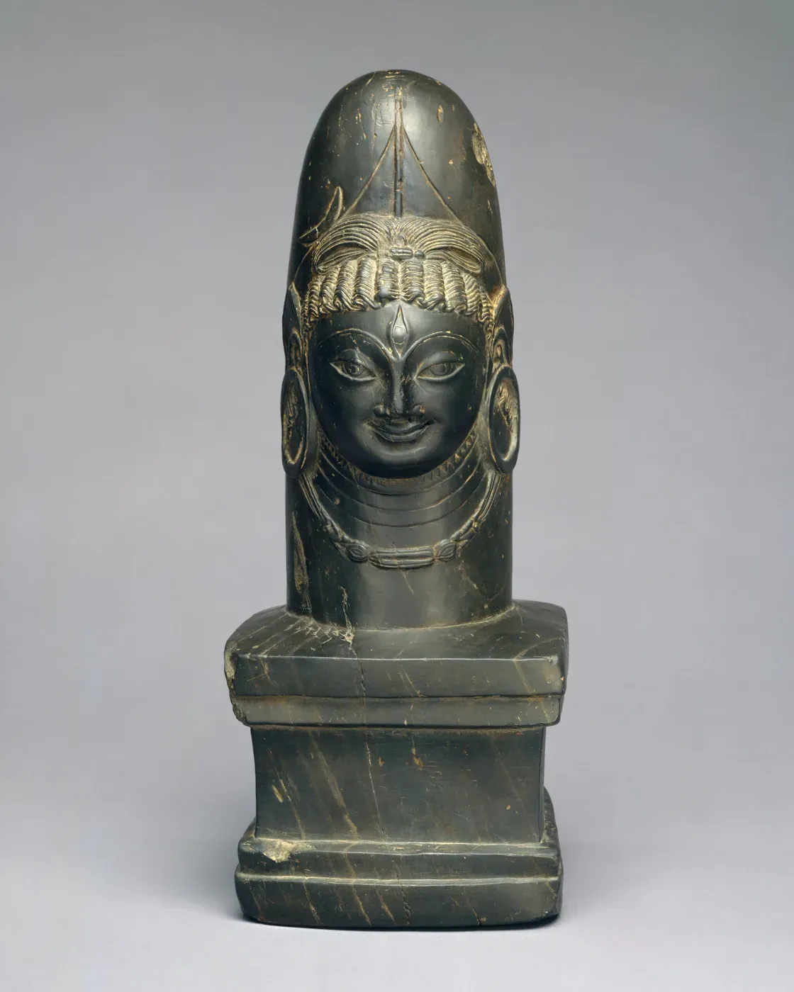 A polished dark stone shaft with a rounded top rising from a square stepped base. A smiling face is carved on the front of the shaft, with large round earrings, a necklace, an eye in the forehead and hair drawn up in loops.