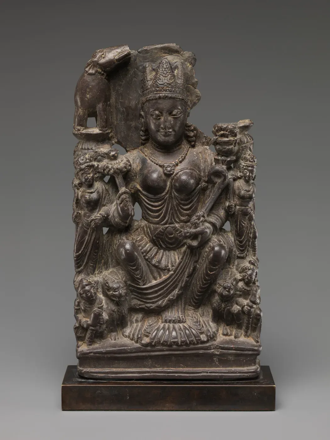 A dark stone carving of a goddess seated on a lotus between two attendants, holding a lotus stalk and a horn-shaped object. An elephant stands on a column above her right shoulder, the matching one on the other side is broken away, and lions sit at her feet.