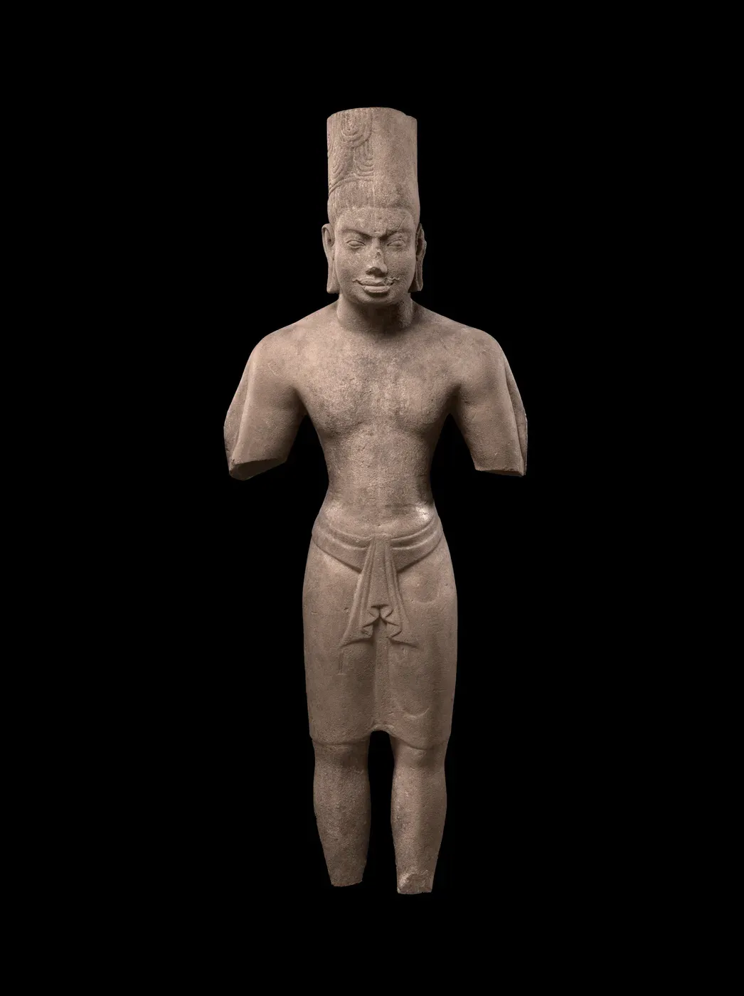 A sandstone figure of a standing man, photographed against black. He wears a tall cylindrical headdress and a short wrapped garment; both arms are broken off above the elbow and the feet are missing.
