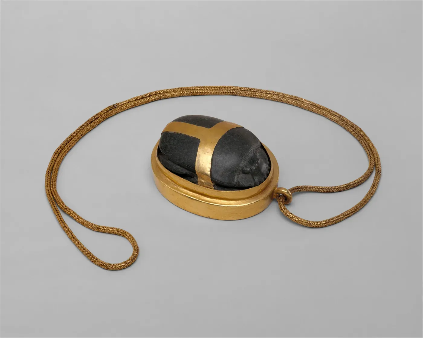 A dark stone scarab beetle set in a gold oval mount, with gold bands crossing its back, attached to a long plaited gold chain that loops around it.