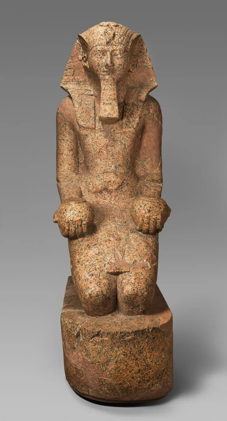 A red granite statue of a kneeling king wearing a striped headcloth and a long false beard, holding a round jar in each hand above the knees, on a rounded base.
