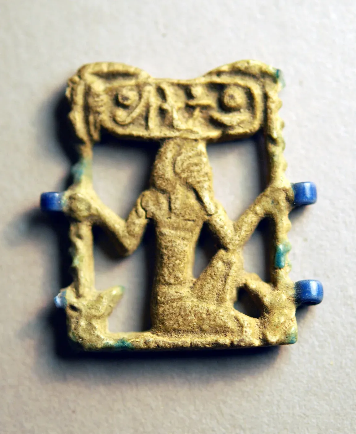 A small openwork faience amulet: a kneeling god with raised arms grasps a notched palm rib on each side, beneath a cartouche with the king's name, with blue loops at the edges.