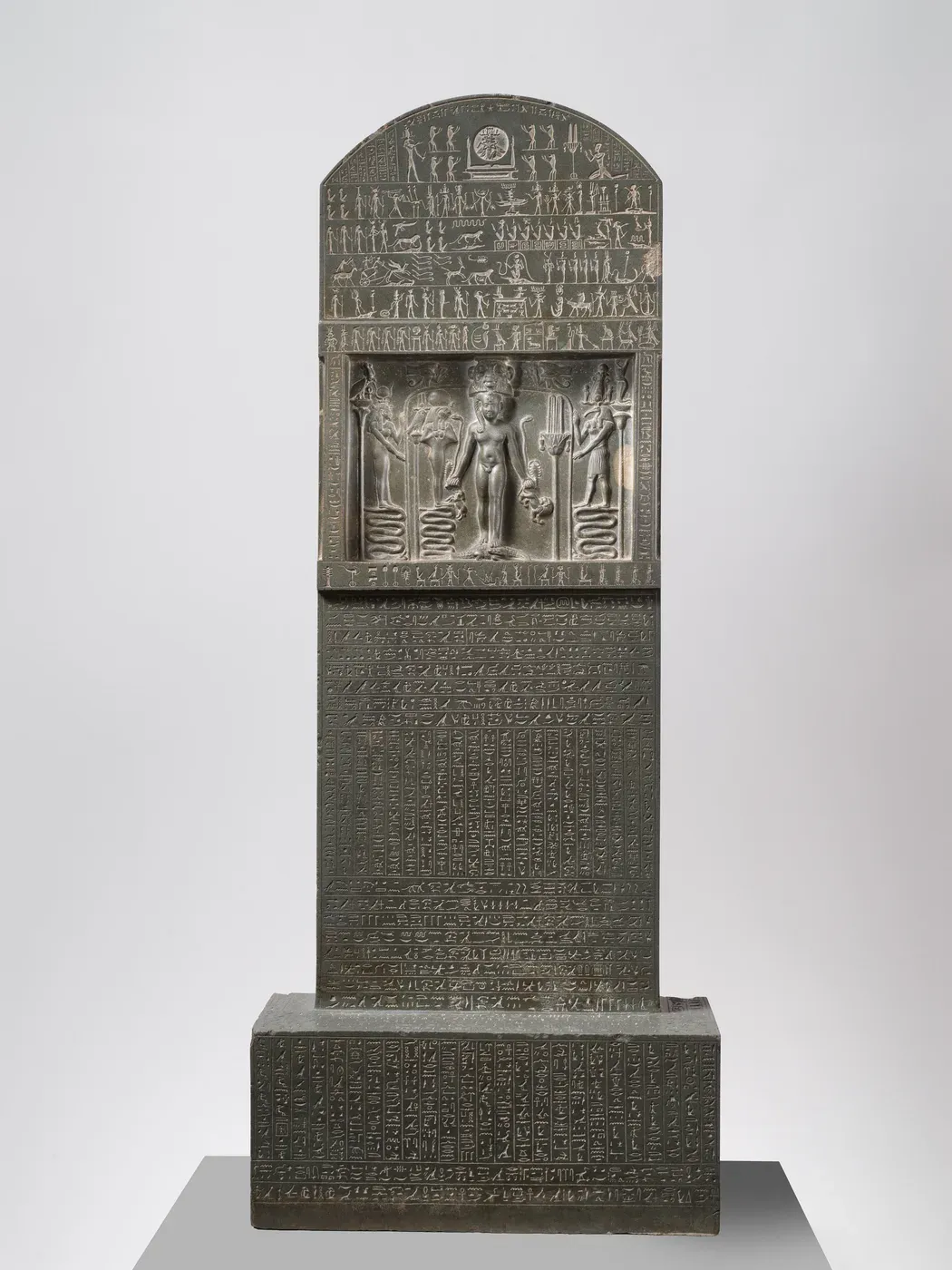 A tall, round-topped slab of dark stone on a block base, covered with rows of small hieroglyphs and tiny figures. In a sunken panel near the top a nude child stands facing forward on two crocodiles, holding snakes and animals, between gods who stand on coiled snakes.