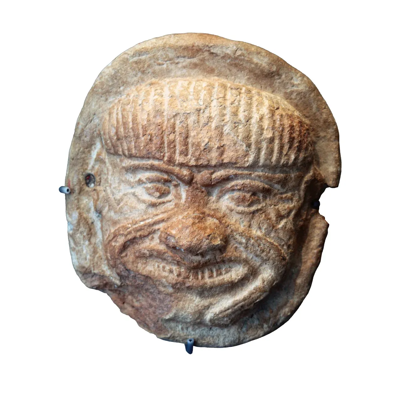 A terracotta head of Humbaba with heavy brows, wide eyes, exposed teeth, and a curling beard.