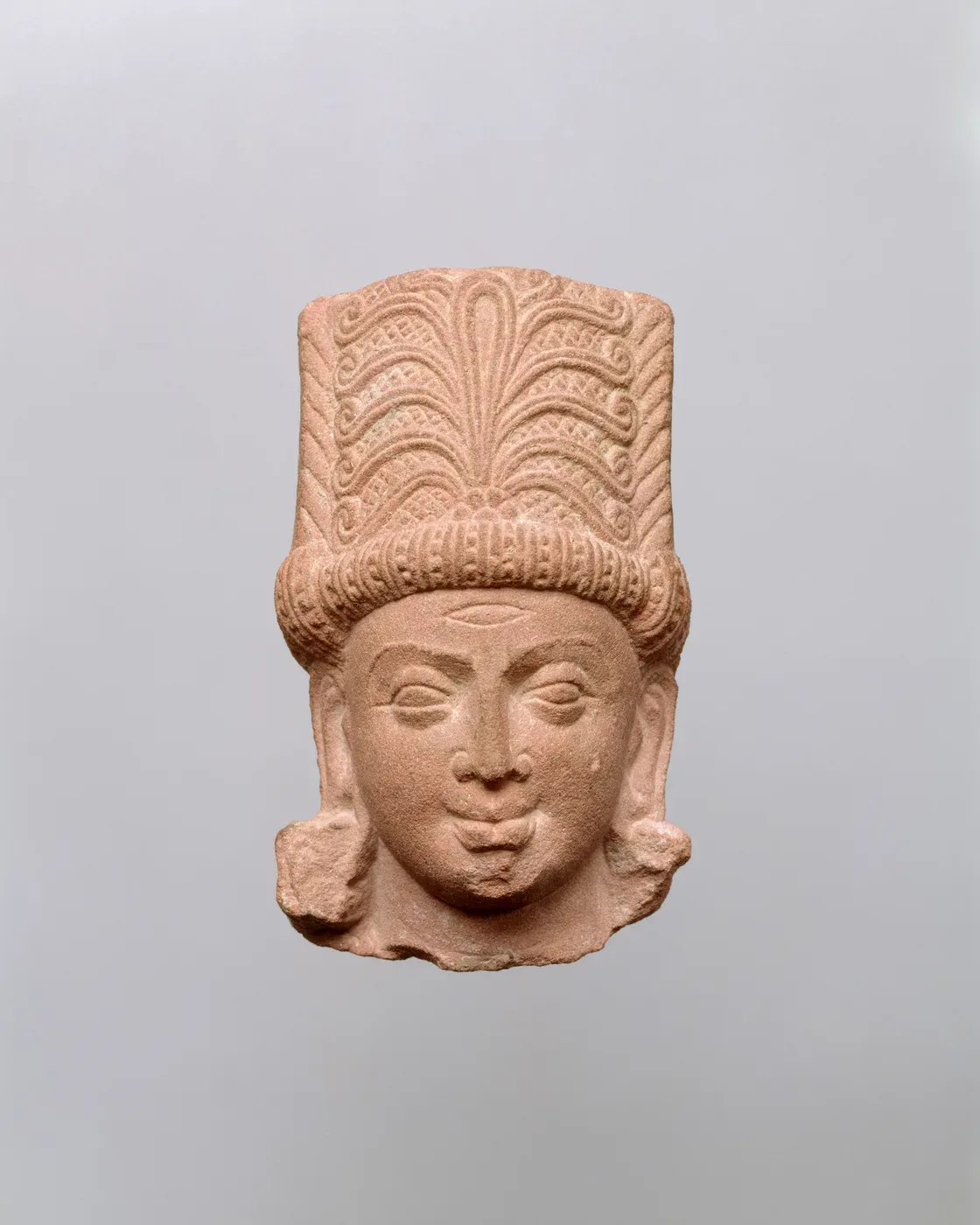 The head of a red sandstone figure. It wears a tall, flat-topped crown carved with a fan-shaped pattern above a thick band, and a small horizontal eye is cut into the forehead.