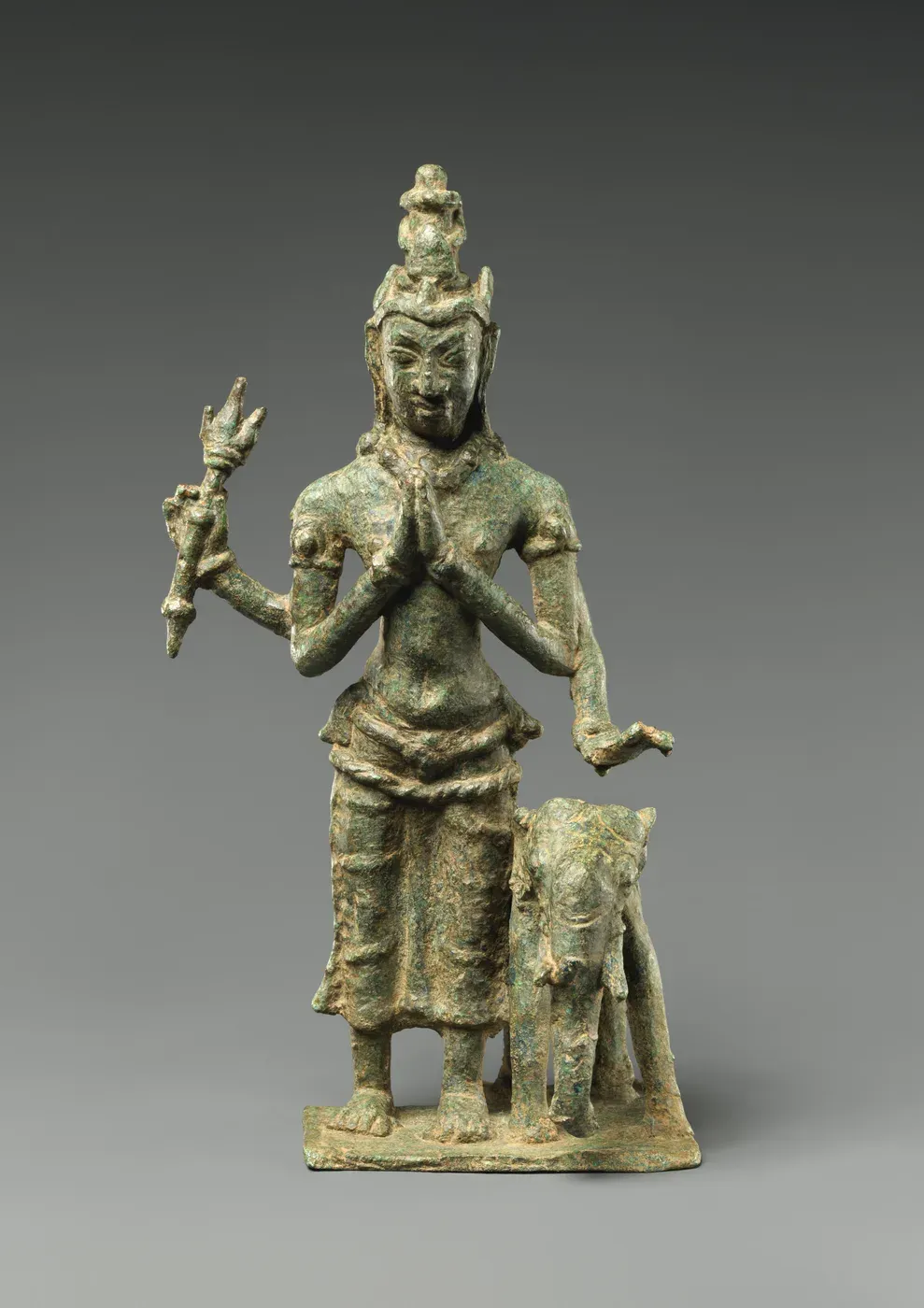 A small green-corroded bronze of a standing crowned figure with four arms. Two hands are pressed together at the chest, one raised hand holds a short ribbed weapon, and a small elephant stands beside his left leg.