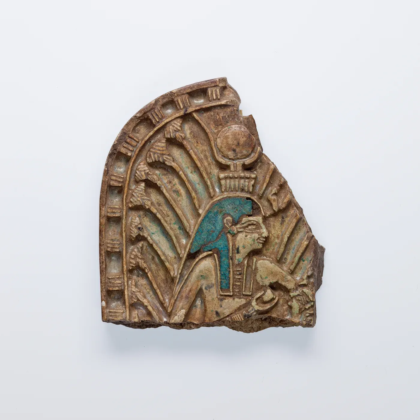 A broken, round-topped plaque of glazed stone carved in relief. A goddess in a blue wig and a crown of cow horns and a sun disk sits among tall papyrus stalks with one hand at her breast; a small head survives at the broken lower edge.