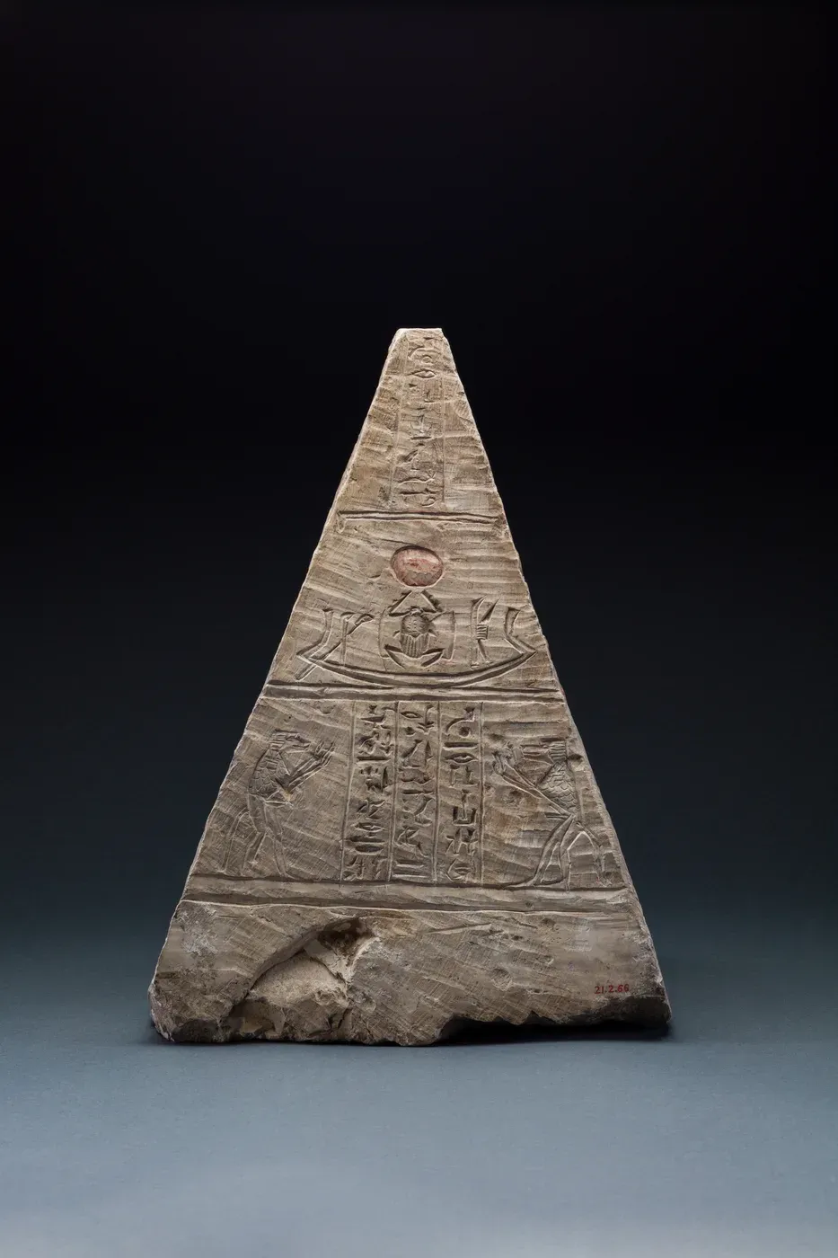 A small limestone pyramid carved on one face with a boat carrying a scarab beetle that holds up the sun disk, above two baboons raising their arms on either side of columns of hieroglyphs.