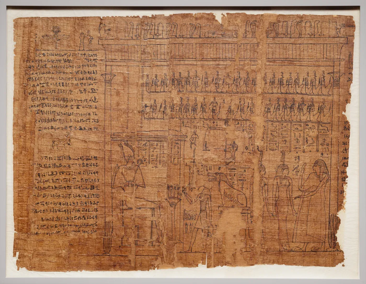 A sheet of brown papyrus drawn in black ink. At the left is a column of script. The rest shows a hall framed by columns: a god sits enthroned in a kiosk at the left, a jackal-headed figure tends a balance in the centre beside a crouching beast on a stand, and at the right a robed person stands with two goddesses who wear feathers on their heads. Two rows of small seated figures fill the space above.