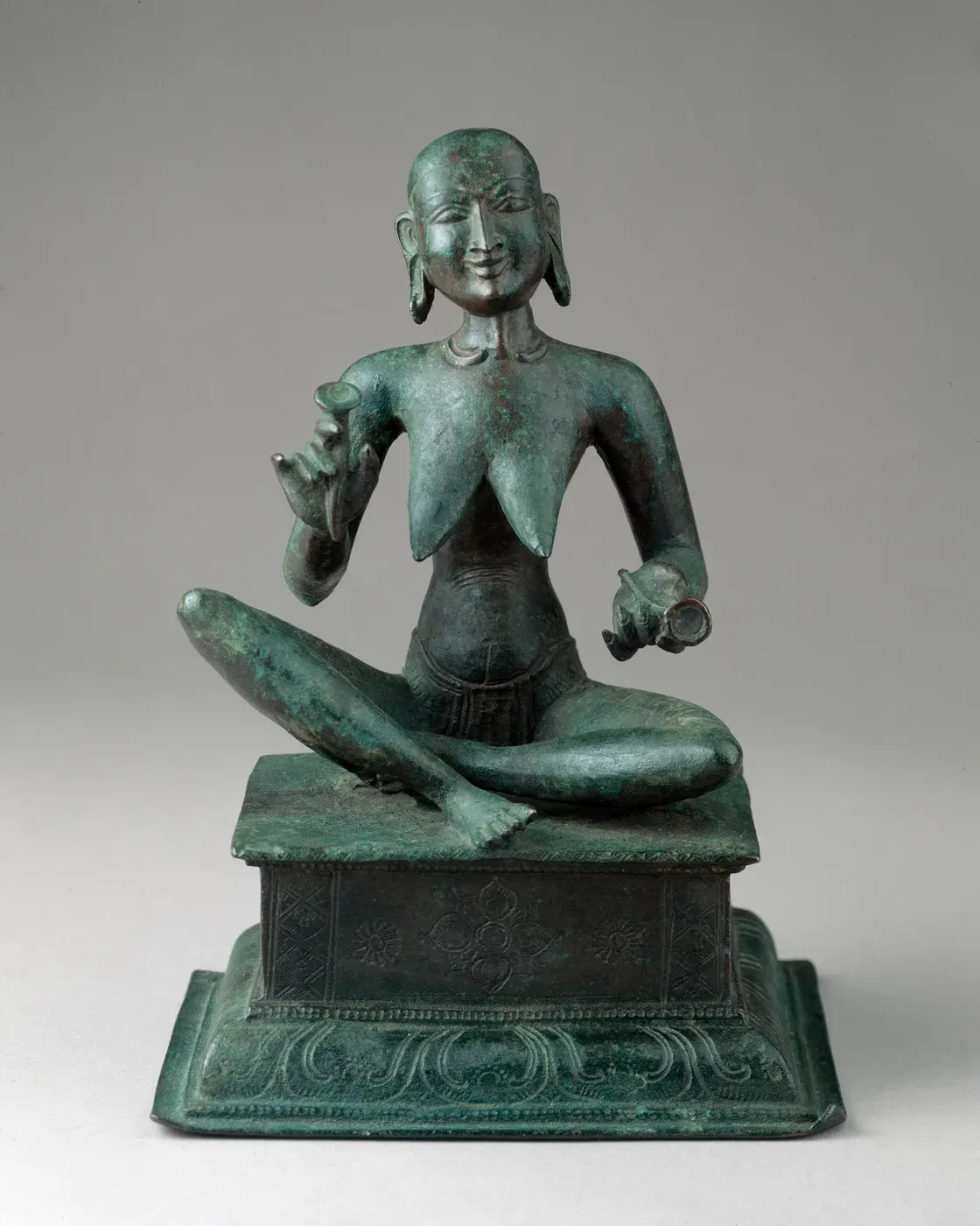 A green bronze of a very thin, smiling woman seated cross-legged on a rectangular base. Her ribs and collarbones show, her head is nearly bald, and she holds a small cymbal in each hand.