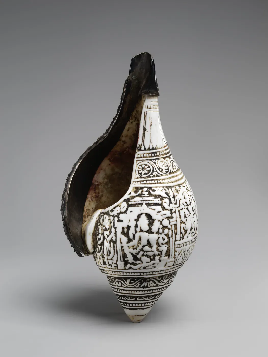 A large white conch shell standing on its point, its surface carved in low relief with bands of ornament and, in the centre, a small seated figure inside a pillared pavilion. The dark inner lip of the shell shows at the left.