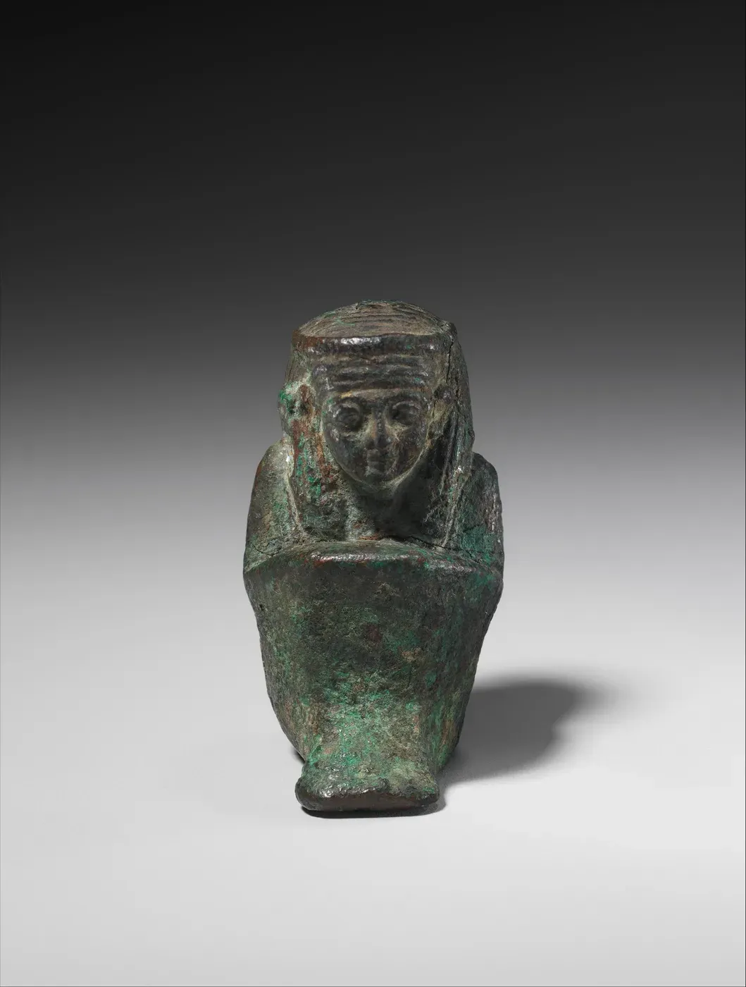 A tiny dark metal figure with green corrosion, seen from the front: a woman seated on the ground with her knees drawn up under a close wrap, wearing a long wig banded at the brow. The top of her head is flat.