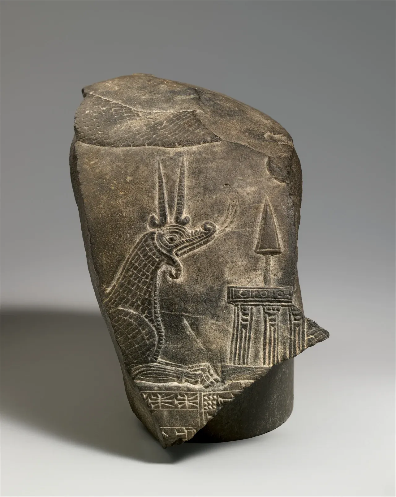 A broken Babylonian limestone monument carved with a long-necked dragon facing Marduk's spade emblem on a raised stand.