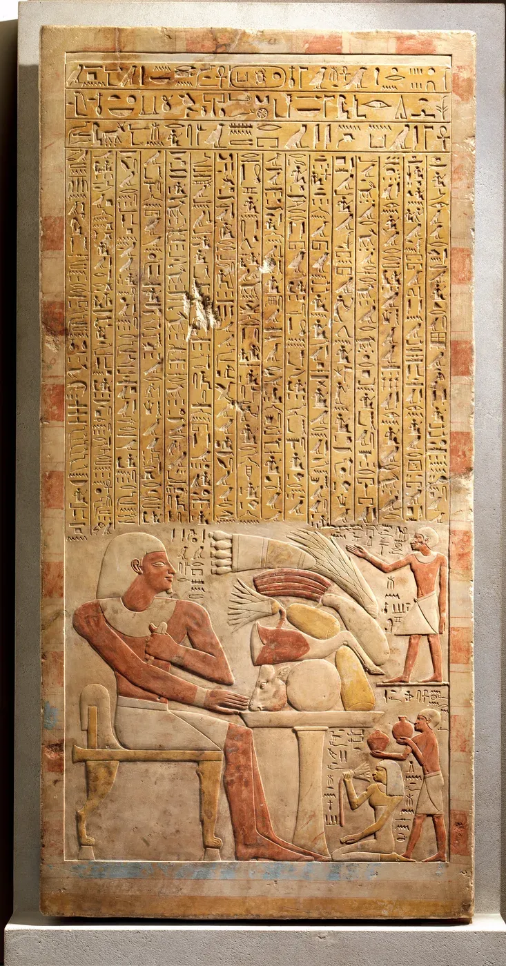 A tall limestone slab. Its upper half is filled with columns of hieroglyphs painted yellow. Below, a man sits on a chair before a table piled with food, while three smaller figures face him with offerings.