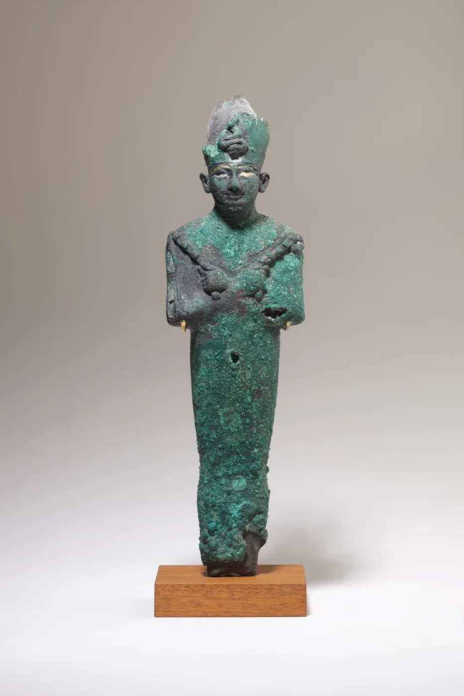 A green-corroded metal figure of Osiris on a modern wooden base: a tightly wrapped body with the hands crossed on the chest holding two sceptres, a faintly smiling face, and a tall crown damaged at the top.