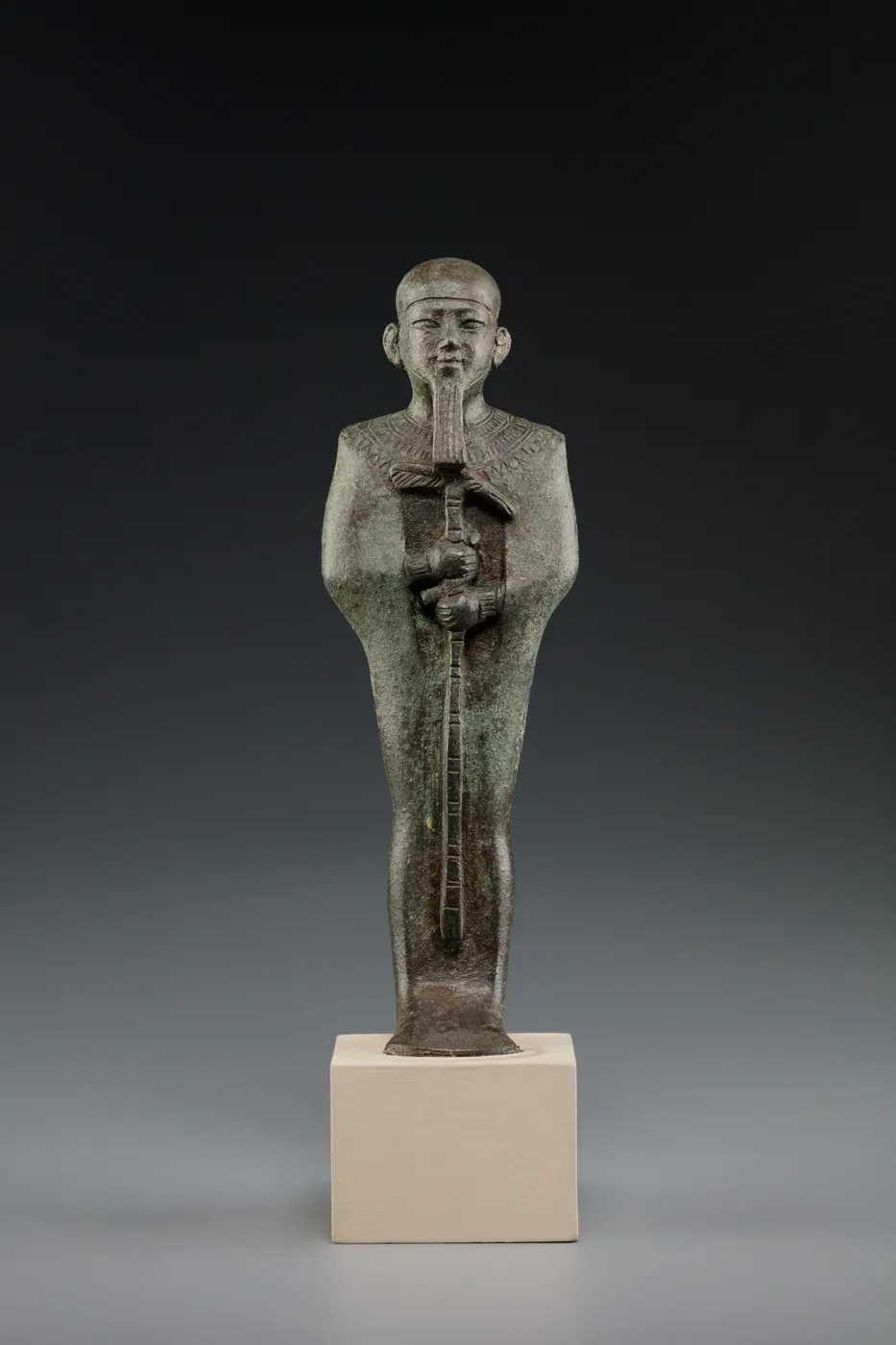 A dark metal figure of Ptah standing on a modern base, wrapped close to the body, wearing a tight cap and a straight beard, and holding a staff upright in both hands.