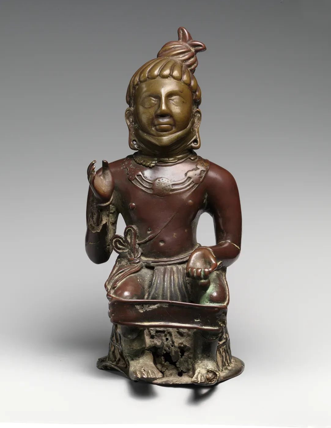 A reddish-brown bronze figure of a man seated with a strap around his knees. His hair is tied in a large topknot, he wears a heavy necklace, his right hand is raised with the palm outward, and his left hand rests on his knee holding a small object.