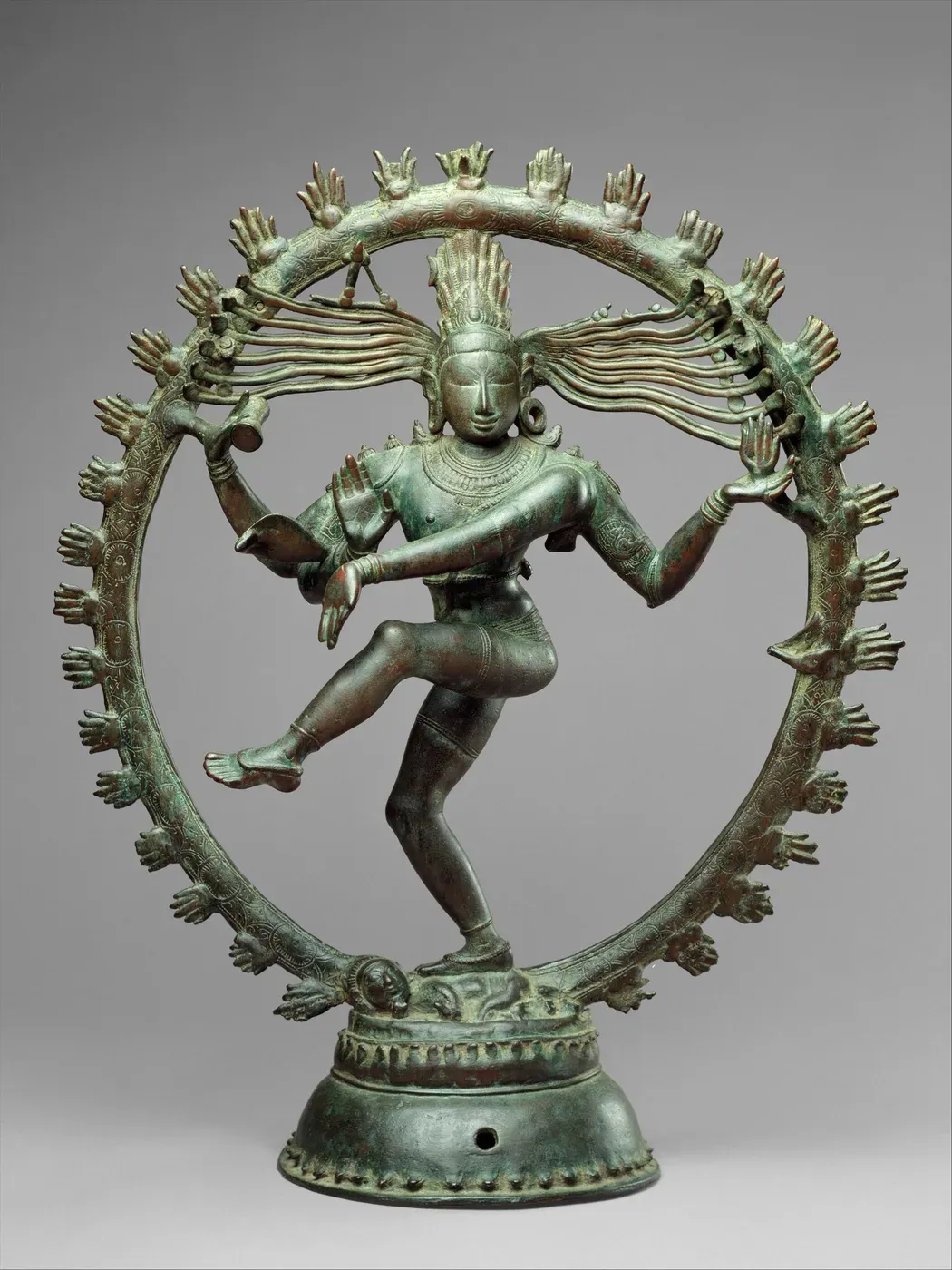 A green-patinated bronze of a four-armed god dancing inside a ring edged with small flames. He balances on one bent leg on a small crouching figure, his other leg lifted across his body, and locks of his hair stream out to both sides.