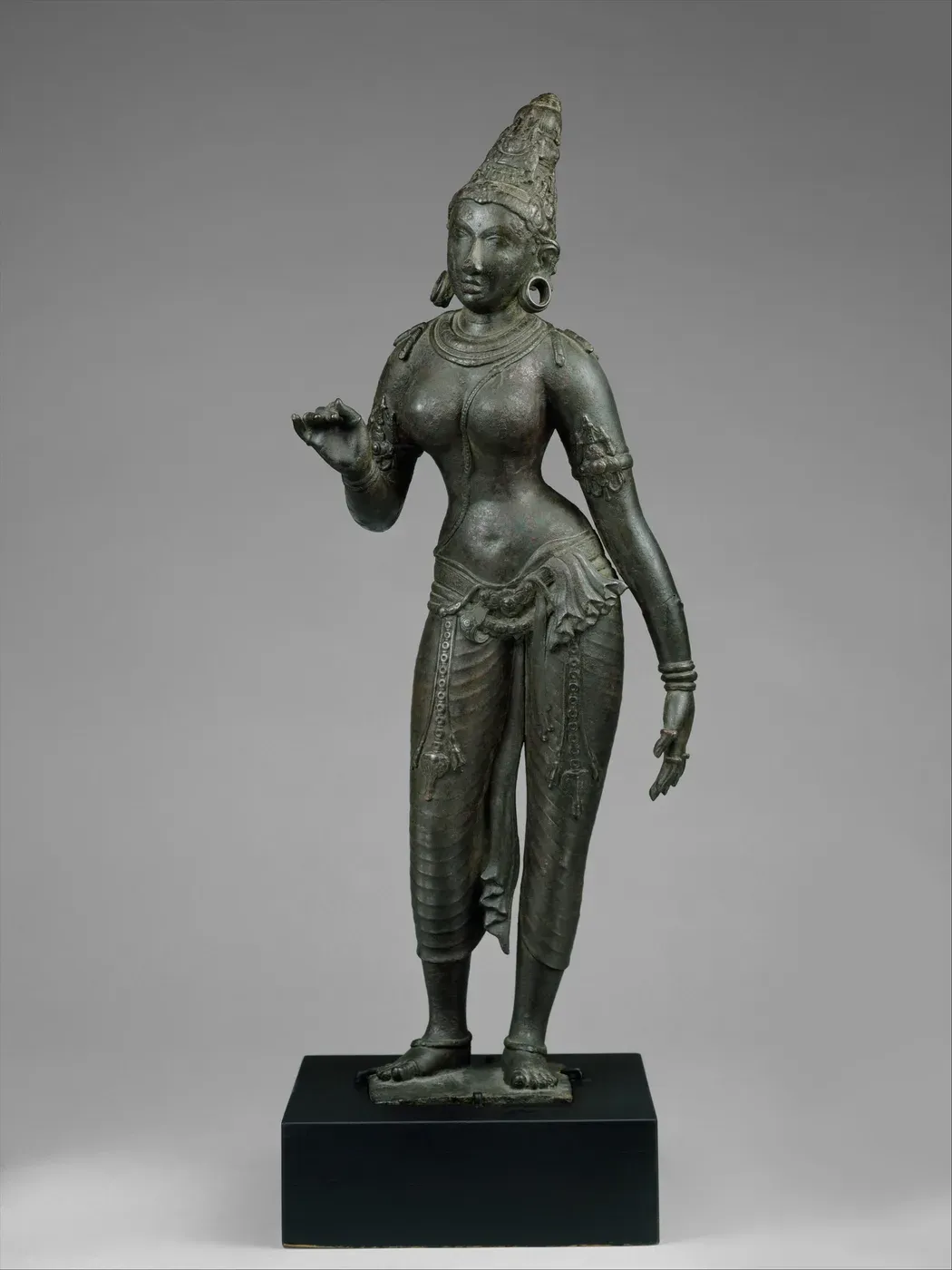 A dark bronze of a slender standing woman on a square block. She wears a tall conical crown and a long patterned lower garment, her weight rests on one hip, and her right hand is raised with the fingers curled as if holding a flower.