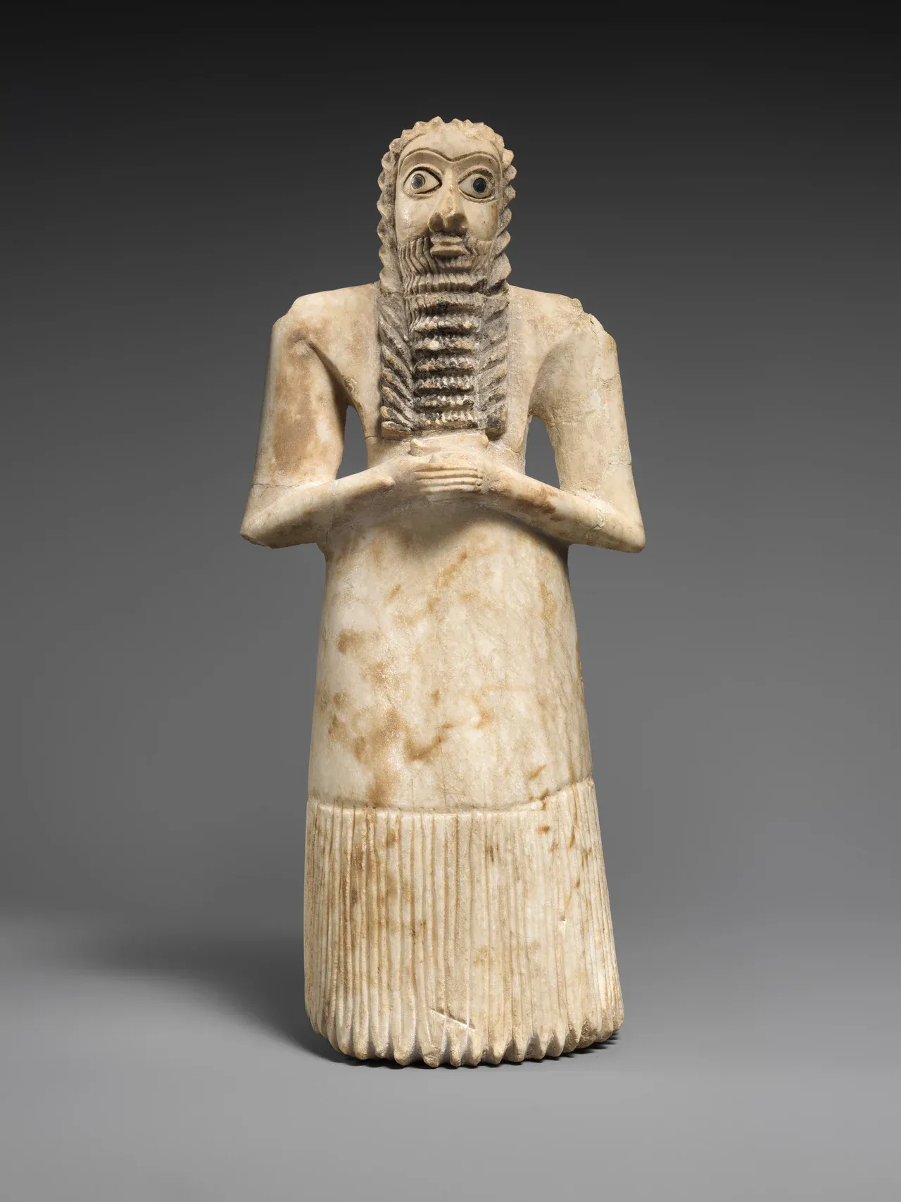 A pale stone figure of a bearded man with large inlaid eyes, clasped hands, and a long fringed skirt.