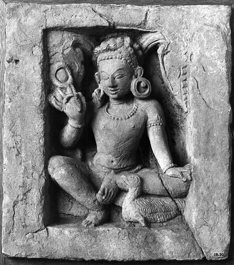 An old black-and-white photograph of a stone relief in a square frame. A figure with large round earrings sits with one knee raised, holding up a looped cord in his right hand, and a long-necked bird sits by his knee.