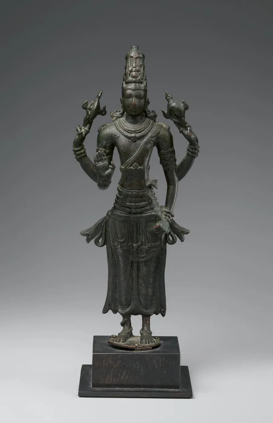 A dark bronze figure of a four-armed god standing straight on a small round base set on a modern block. He wears a tall crown and a long lower garment; his two raised hands each hold a small object on a flame-like stand, one lower hand is raised with the palm outward and the other rests at his hip.