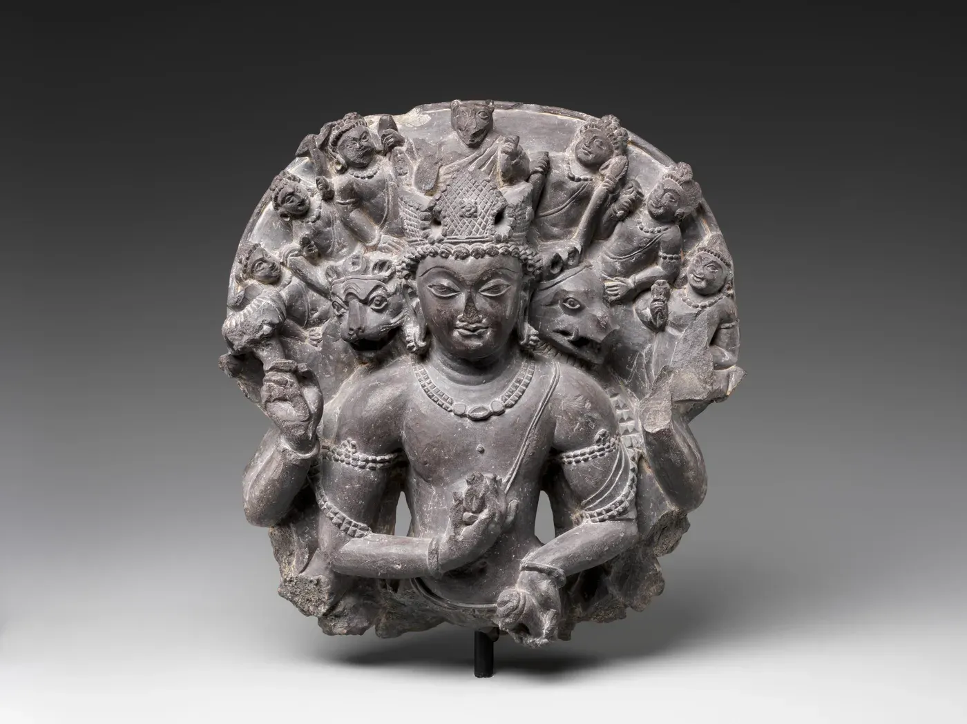 A fragment of dark stone carved with the upper body of a god with several arms. Beside his crowned human face are the heads of a lion and a boar, and behind him a round halo is crowded with many small figures.