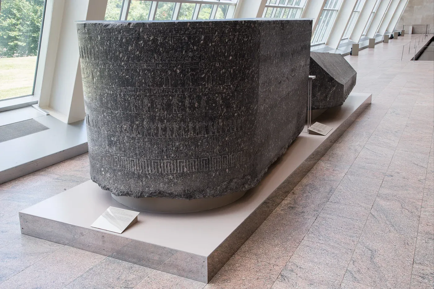 The rounded end of a massive dark stone sarcophagus on a platform in a museum gallery. Its surface is carved in horizontal bands of small figures and hieroglyphs. The detached lid lies behind it.